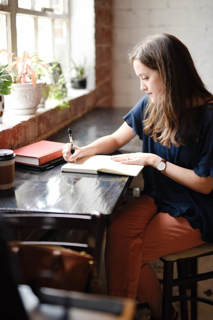 A young woman writing in a notebook at a wooden table with plants and books nearby.