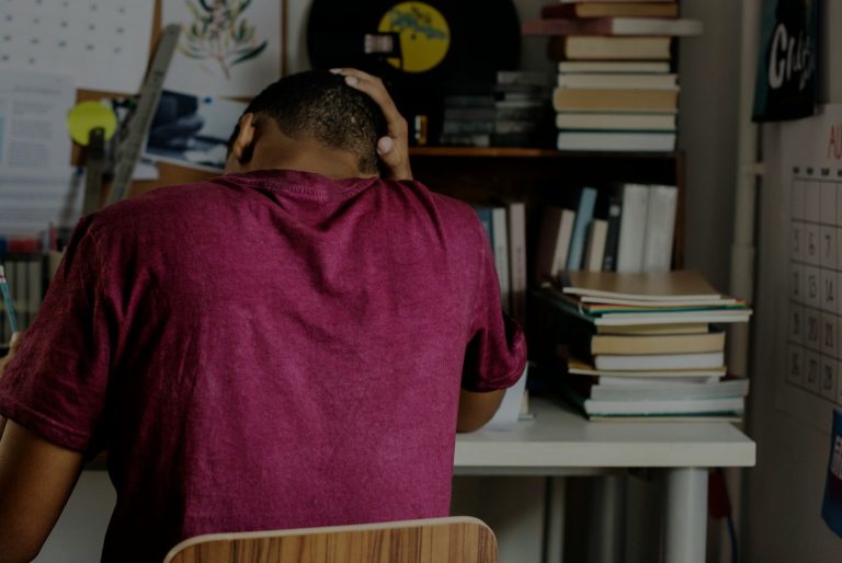 Teenager working hard A person in a burgundy shirt sitting at a desk, looking down, surrounded by books.