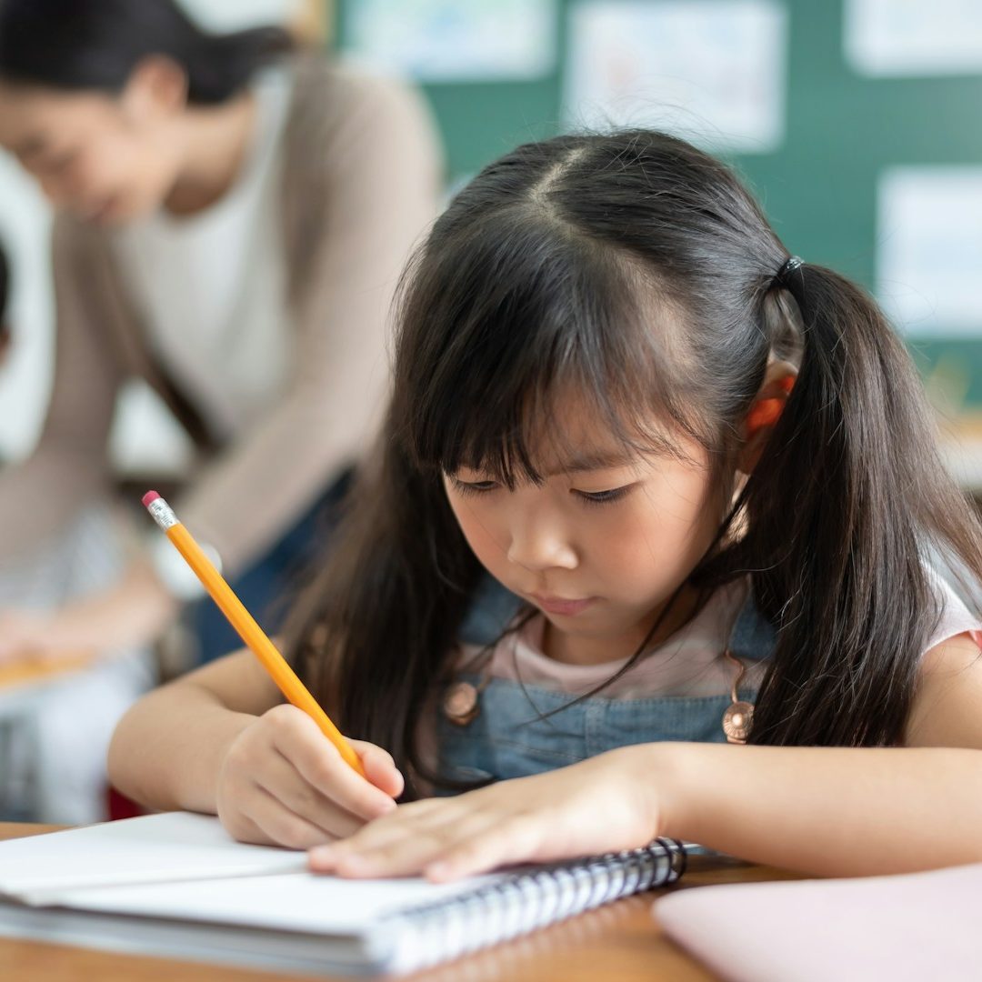 A young girl focused on writing in a notebook at a classroom desk.