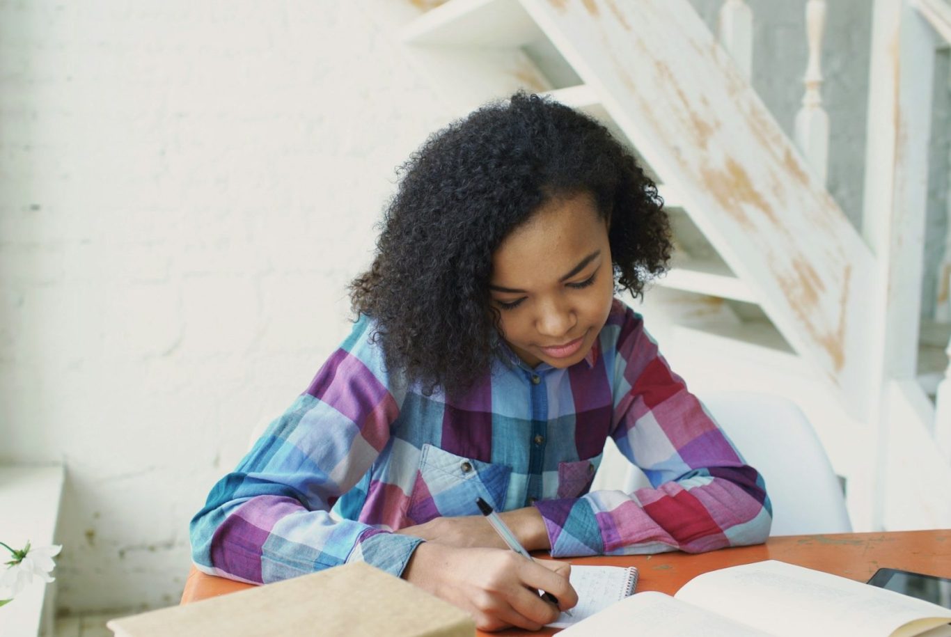 Teenager working at table Young girl with curly hair focused on writing at a table, surrounded by a bright room.