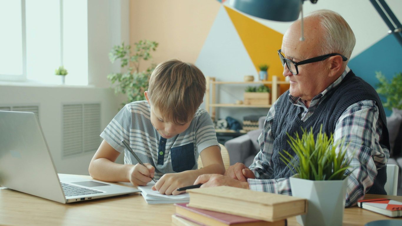 Boy and man working A boy studies at a table while an elderly man watches him, both engaged in learning.