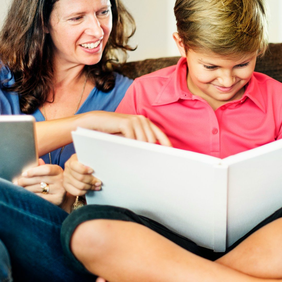 A woman and a boy are smiling together while reading a book on a sofa.