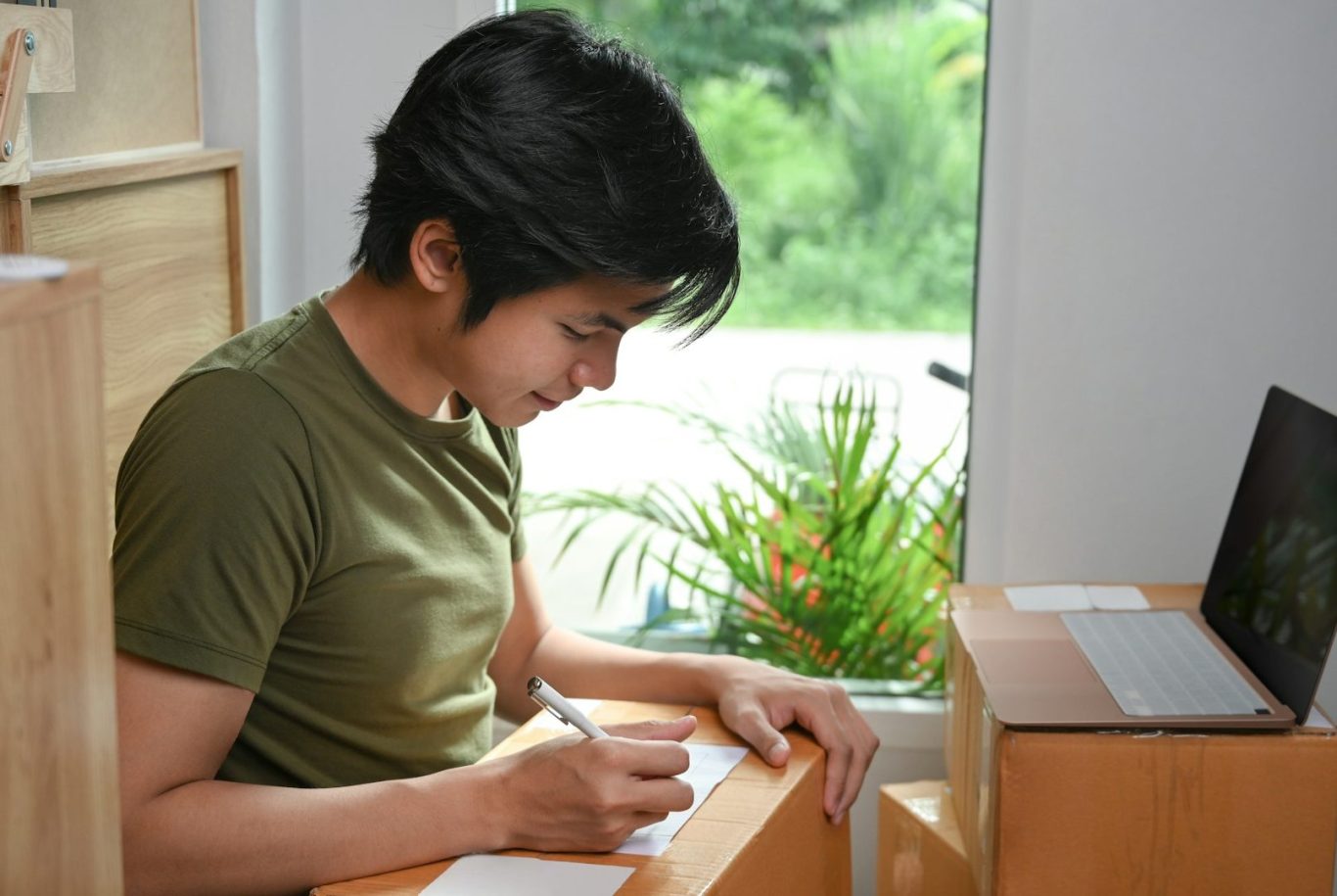 A person writing at a desk by a window with greenery outside.